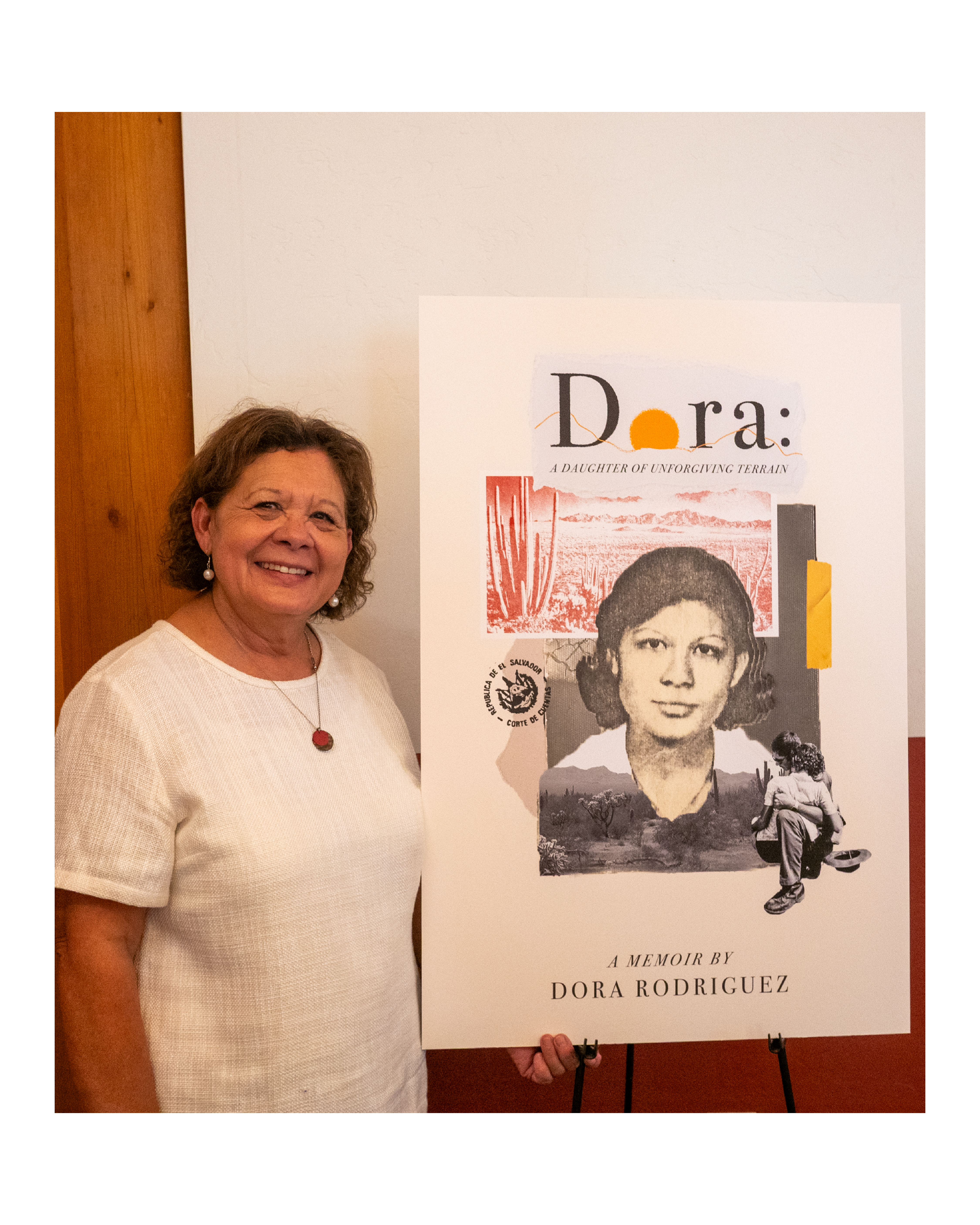Dora Rodriguez at a book event, standing beside a poster of her memoir, Dora: A Daughter of Unforgiving Terrain.